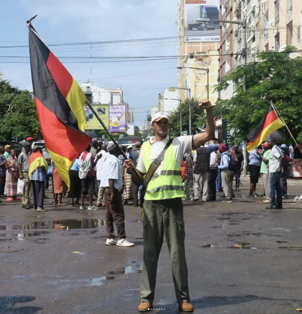 Demonstration ehemaliger DDR Arbeitnehmer in Maputo, ein Mann in Warnweste, dahinter deutsche und mosambikanische Fahnen