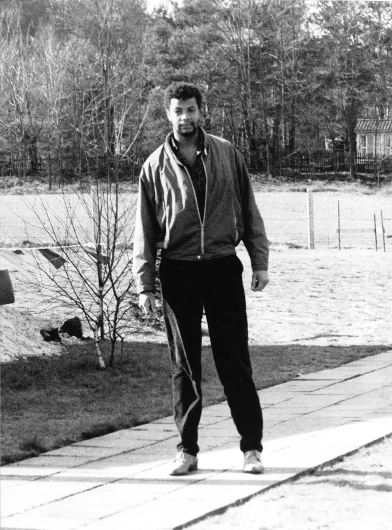 Arnaldo Soares Mendes in heller Jacke auf einem Plattenweg vor einer Wiese und Kiefern, Eberswalde, späte 1980er Jahre