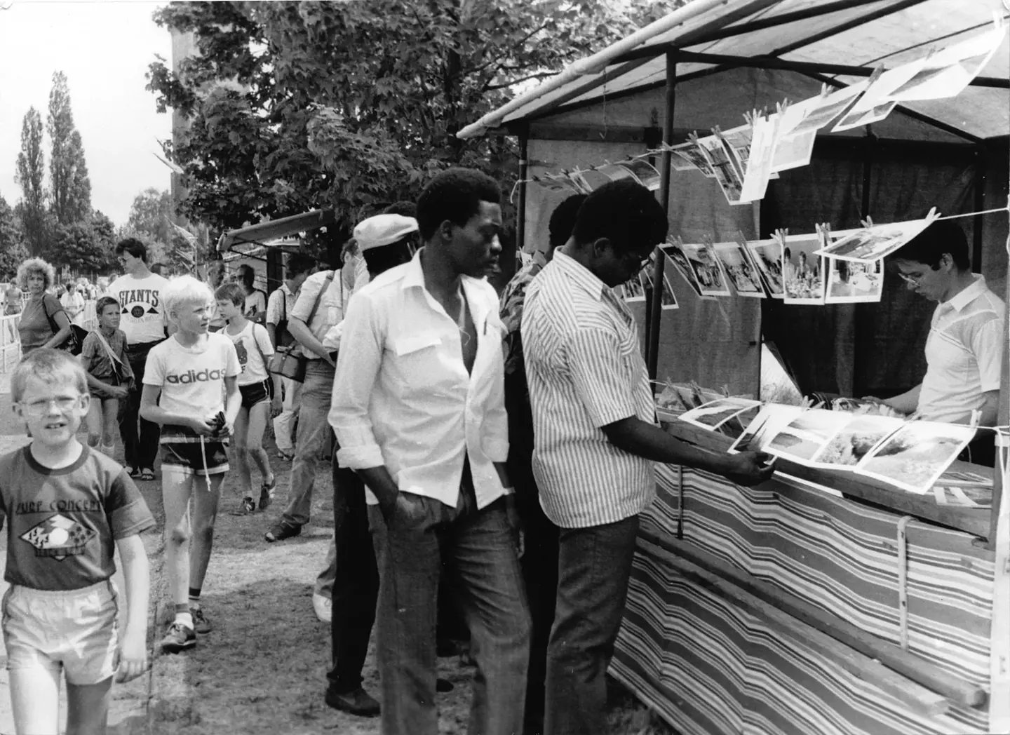 Mosambikanische Männer betrachten an einem Stand ausgehängte Fotografien, dahinter Kinder und Besucher eines Festes unter Bäumen.